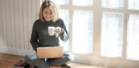 a girl is sitting with a laptop and a cup of coffe in her hands